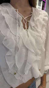 Top blanco de manga larga, confeccionado en tejido ligero y traslúcido. Presenta escote en V con cordones entrelazados y volados decorativos en el frente. Las mangas son largas y abullonadas con puños ajustados.
