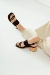 Sandalias de gamuza color marrón con taco cuadrado de madera. Presentan dos tiras anchas, una superior lisa y una inferior decorada con ojales metálicos plateados. Cuentan con cierre de pulsera con hebilla ajustable en el tobillo.