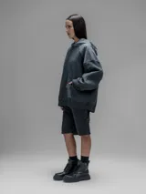 Hoodie oversize de color gris oscuro, confeccionado en frisa de algodón con recortes en textiles técnicos. Presenta capucha forrada, bolsillo frontal tipo canguro y un bolsillo adicional con solapa en la parte inferior.