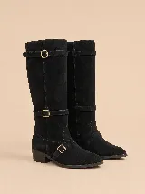 Bota alta de gamuza color negro, con caña ancha, taco bajo cuadrado y punta redondeada. Cuenta con tres tiras con hebillas metálicas doradas ajustables alrededor de la caña, el tobillo y el empeine.