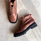 Botas de cuero color marrón con suela track negra gruesa, cordones naranjas y cierre frontal con cremallera.