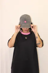Gorra tipo baseball de color gris con efecto desgastado, presenta un detalle circular en color rosa y verde en la parte frontal.