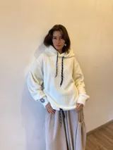 Buzo tipo hoodie de tela de peluche color manteca, con capucha y cordones ajustables. Presenta mangas largas con puños elásticos y una insignia bordada de una leona en uno de los puños.