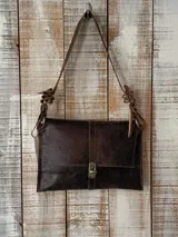 Cartera de cuero marrón con textura desgastada, diseño rectangular con solapa frontal y cierre metálico a presión. Presenta una correa de hombro única con detalles decorativos de cuero trenzado y anillas metálicas.