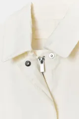 Gabardina oversize color blanco roto, con cuello solapa, manga larga y bolsillos laterales ocultos. Presenta cintura ajustable con cordones a tono y forro interior.