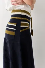 Falda midi de lana en color marrón, con diseño acampanado y tejido acanalado. Presenta una cintura elástica con cordón de ajuste, detalles de rayas en tonos beige y blanco en la parte superior, y costuras decorativas en el ruedo.