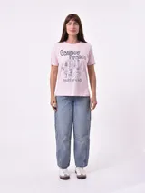 Remera de manga corta color beige claro con cuello redondo. Presenta un estampado gráfico central con un plato de pasta rodeado por un borde azul y blanco, sobre un fondo de franjas verticales rojas y blancas. El estampado incluye texto en cursiva en tonos turquesa y rojo.