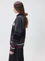 Pullover de punto con textura aterciopelada, diseño oversize, cuello polo con cierre frontal parcial y detalles de pespuntes en contraste. Presenta rayas horizontales en tonos tierra y un parche bordado con la letra M en el pecho.