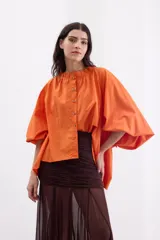 Camisa holgada de popelina de algodón con rayas verticales en tonos rosado, gris y blanco. Presenta cuello alto con volado, hombros caídos, mangas anchas abullonadas y cierre frontal con botones.