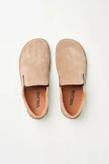 Zapatillas tipo slip-on de gamuza color beige, con diseño barefoot y suela de corcho texturizado. Presenta elásticos laterales negros para un calce fácil.