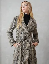 Trench de ecocuero con estampado animal print de serpiente en tonos beige, marrón y negro. Presenta cuello con solapa ancha, doble abotonadura frontal con botones negros, cinturón ajustable con hebilla metálica, mangas largas con presillas y botones en los puños, canesú suelto en la espalda, bolsillos laterales y forro interno.