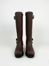 Bota de caña alta estilo biker, confeccionada en cuero negro. Presenta tres hebillas metálicas decorativas, cierre lateral y taco de madera.