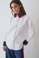 Camisa de lino blanca con cuello y puños tejidos a mano en crochet azul con borde rojo. Presenta cierre frontal con botones de madera, bolsillo frontal y costuras en contraste.
