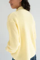 Saco tejido de punto color amarillo claro, con cuello alto asimétrico abotonado, botones oscuros contrastantes en la parte frontal y mangas largas con puños ajustados.