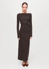 Vestido largo de corte ajustado en color marrón oscuro, con escote bote y mangas largas. Presenta un diseño minimalista con un sutil fruncido lateral en la cintura que realza la silueta.
