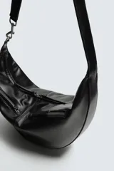 Bolso de hombro de diseño curvo en forma de media luna, confeccionado en material sintético negro. Presenta dos bolsillos frontales con cierre de cremallera y un compartimento principal con cierre. Incluye un asa de hombro integrada y detalles de costuras visibles.