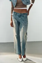 Jeans rectos de tiro medio en denim azul con efecto deslavado. Presentan bandas laterales con detalles brillantes, bolsillos delanteros y traseros, y aberturas en los tobillos con botonadura metálica.
