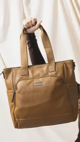 Cartera tipo tote marrón de diseño práctico y versátil, confeccionada en símil cuero con textura granulada. Cuenta con asas cortas fijas, bolsillos exteriores con cierre al frente y atrás, y bolsillos laterales abiertos. Su interior es amplio e incluye bolsillos organizadores. Viene con una correa ajustable y desmontable.