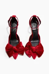 Zapatos de tacón tipo pump en terciopelo rojo, con punta fina y un lazo grande decorativo en el empeine. Presentan una pulsera ajustable al tobillo con cierre de hebilla y tacón fino forrado.