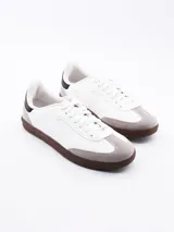 Zapatilla estilo urbano de color blanco, con refuerzo de gamuza gris en la puntera, suela de goma marrón y detalles en negro en el talón.