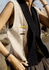 Chaleco abierto de tejido de punto, con diseño de bloques de color verticales en gris claro, negro y gris oscuro. Presenta un cuello ancho tipo chal y un detalle de tres pequeñas aplicaciones bordadas horizontales en tonos verde oliva, naranja quemado y celeste pálido en el frente.