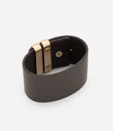 Pulsera ancha de material sintético color marrón oscuro, con diseño de cinta larga y dos tiras metálicas doradas aplicadas en el centro. Cuenta con un cierre de pino ajustable.