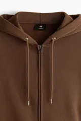 Campera con capucha H&M de corte oversized, confeccionada en tejido pesado de mezcla de algodón con interior cepillado. Presenta cierre frontal, hombros caídos, capucha forrada con cordón de ajuste y bolsillos tipo canguro. Puños y bajo acanalados.