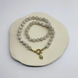 Collar de cuentas de piedra de la luna de 8 mm, con nudos de hilo color caramelo entre cada cuenta. Presenta un cierre frontal de argolla tipo mosquetón y un dije en forma de corazón con baño de oro.