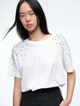 Remera blanca de corte oversize con cuello redondo y mangas cortas. Presenta apliques de strass o piedras brillantes distribuidos en la zona de los hombros.