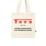 Tote bag color beige de lona, con diseño gráfico estampado en rojo y negro que incluye la inscripción "Toto Since 1934" y "Donde empezaron tus primeros pasos". Cuenta con dos asas superiores de hombro.