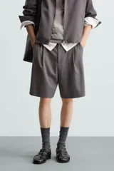 Bermuda de corte relajado color negro, con pliegues frontales, cierre de cremallera y botón, bolsillos laterales y bolsillos traseros.