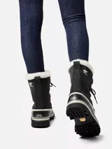 Bota de nieve Sorel modelo Caribou, con diseño impermeable de caña media. Presenta una parte superior de nubuck negro con costuras selladas, cordones frontales y una base de goma vulcanizada resistente. Incluye un forro interior de fieltro y detalle de pelo sintético en el borde superior.