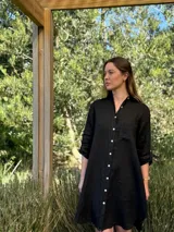 Vestido camisero negro de lino, con cuello camisero, cierre frontal con botones blancos, manga 3/4 con trabillas y bolsillo de parche en el pecho. Diseño asimétrico, más largo en la parte trasera.