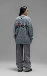 Sweater de punto texturizado color gris, con cuello redondo y diseño oversize. Presenta un bordado en relieve en la espalda con la frase 'art SUPPORT' en letras rojas y negras.