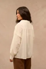 Blusa color crema de manga larga con detalles de alforzas verticales en el frente y hombros, escote en V con cuello mao y puños abotonados.