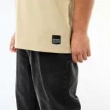 Remera de algodón color beige con cuello en V y manga corta. Presenta un corte holgado y una etiqueta decorativa pequeña en el borde inferior.