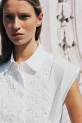 Camisa blanca de popelín con cuello solapa, manga sisa y detalle de guipur floral en el frente. Incluye un cinturón fino del mismo tejido que se ata en la cintura.