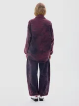 Camisa oversized de denim con lavado acid wash en tonos morados y azul oscuro. Presenta cuello camisero, escote en V, dos bolsillos frontales con tapa y botones oscuros. Las mangas son largas tipo balloon con puño ajustado. El bajo es irregular.