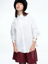 Camisa blanca de corte holgado con cuello clásico y cierre frontal de botones. Presenta un diseño distintivo en los puños con aberturas decoradas con pedrería brillante.