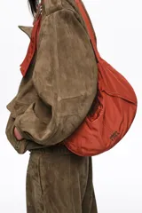 Bolso tipo hobo de la marca Bimba y Lola, modelo Moon mediano, de color rojo óxido. Confeccionado en nylon con diseño de media luna, cierre superior con cremallera, correa ajustable con hebilla metálica y el logotipo de la marca impreso en la parte frontal.