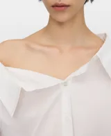 Camisa blanca de algodón con diseño desestructurado, escote asimétrico que deja un hombro al descubierto, manga larga y botonadura frontal.