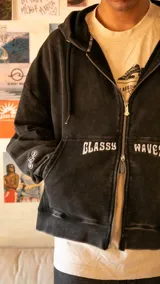 Campera con capucha y cierre frontal doble, confeccionada en algodón con efecto de lavado desgastado color negro. Presenta bordado frontal con el texto 'CLASSY WAVES' y detalle bordado en la manga.
