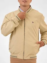 Campera bomber reversible azul marino y beige con cuello, puños y ruedo acanalados con rayas doradas. Tiene cierre frontal y logo bordado en el pecho.
