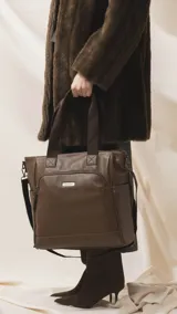 Cartera tipo tote marrón de diseño práctico y versátil, confeccionada en símil cuero con textura granulada. Cuenta con asas cortas fijas, bolsillos exteriores con cierre al frente y atrás, y bolsillos laterales abiertos. Su interior es amplio e incluye bolsillos organizadores. Viene con una correa ajustable y desmontable.