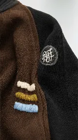 Polera de cuello alto confeccionada en lana lambswool, con diseño bicolor en negro y marrón. Presenta mangas largas con franjas verticales en gris y un parche circular bordado en la manga derecha.