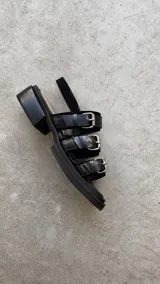 Sandalias de cuero negro con punta semicuadrada, taco cuadrado bajo y tres tiras frontales con hebillas metálicas plateadas. Cuentan con tira trasera con cierre ajustable.