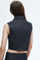 Chaleco corto de denim de algodón rígido, color azul oscuro, con cuello, cierre frontal de botones de metal y dos bolsillos superiores con solapa y botón. Presenta un corte ajustado con canesú y costuras visibles que aportan estructura.