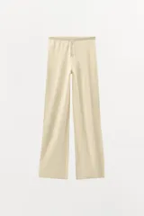 Pantalón de tejido de punto color beige claro, tiro alto, con cintura elástica y detalle de botones frontales. Presenta pernera recta y corte holgado.