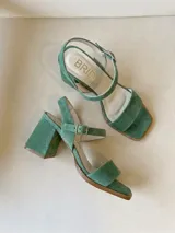 Sandalias de tacón medio ancho y puntera cuadrada, confeccionadas en gamuza color verde pistacho. Cuentan con tira delantera ancha, tira al tobillo con hebilla metálica dorada y talón cerrado.