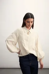 Camisa a rayas verticales color beige y blanco, con cuello clásico, cierre frontal con botón dorado, mangas largas abullonadas con puños elásticos, dos bolsillos frontales y corte holgado.